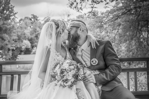 Ein Kuss für die Ewigkeit: Brautpaar im Glück Braut und Bräutigam küssen sich zärtlich auf einer hölzernen Bank im Freien. Der Bräutigam umarmt die Braut, die einen Brautstrauß hält. Alles in Schwarz-Weiß.